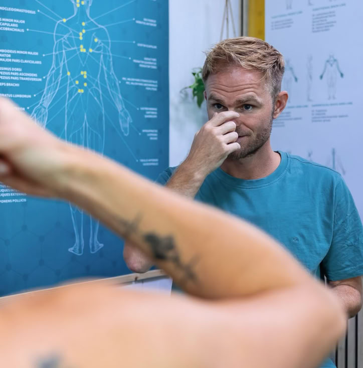 Een man in een blauwe shirt staat in een behandelkamker van de praktijk en knijpt in zijn neus. Op de achtergrond is een poster met anatomische illustraties zichtbaar.