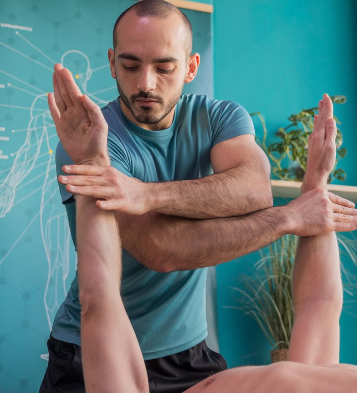 Man in blauwe shirt voert fysiotherapie uit op persoon die op tafel ligt, met armen omhoog.