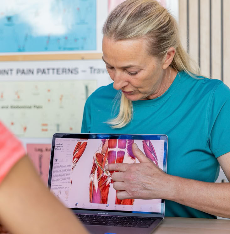 Vrouw in blauwe trui wijst naar anatomische afbeelding op laptop, met posters op de achtergrond.